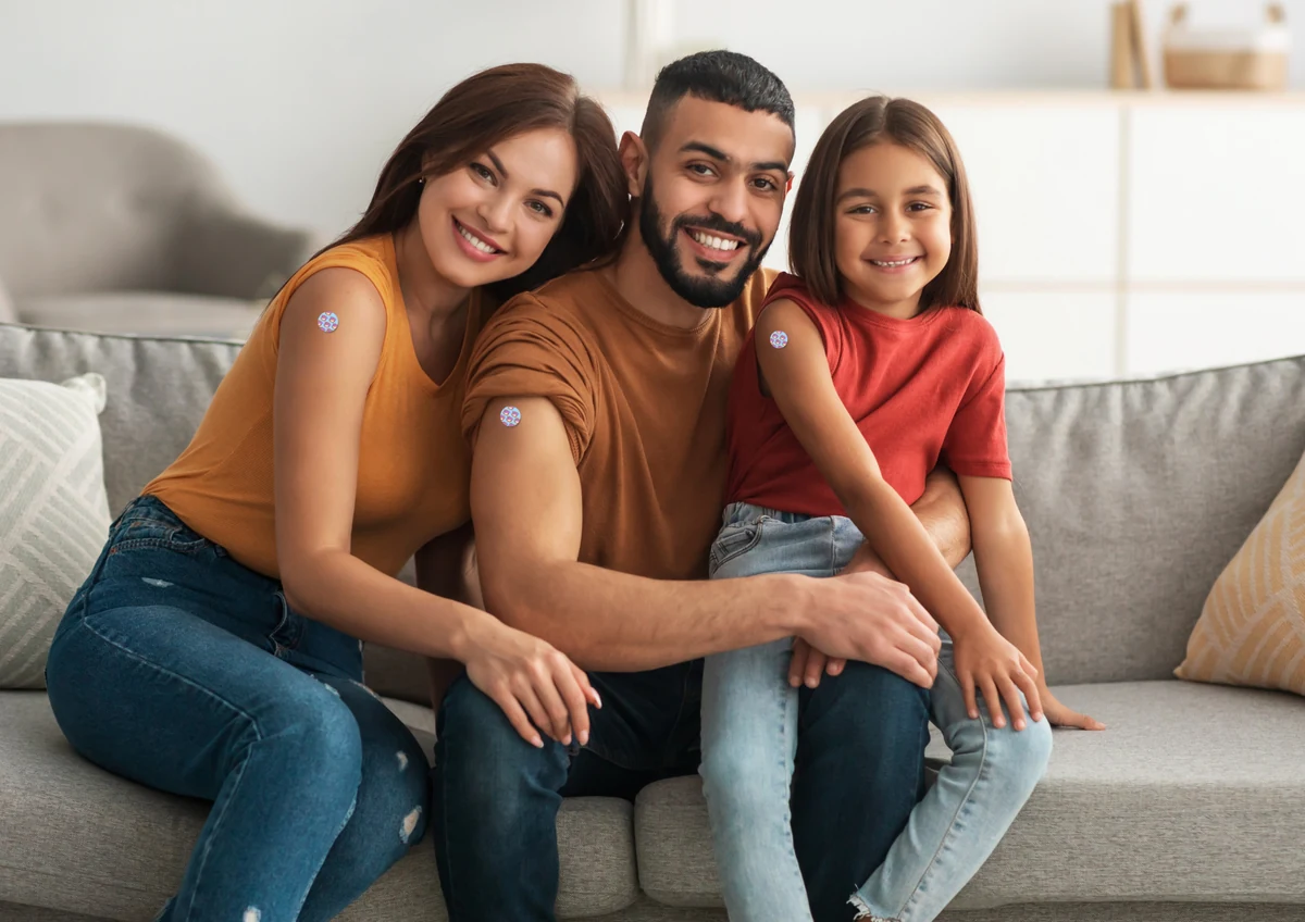 Família sorridente no sofá com os curativos da vacinação: mãe, pai e filha